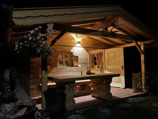 Holzveranda mit Tisch und Stühlen, sanft beleuchtet, umgeben von Pflanzen.