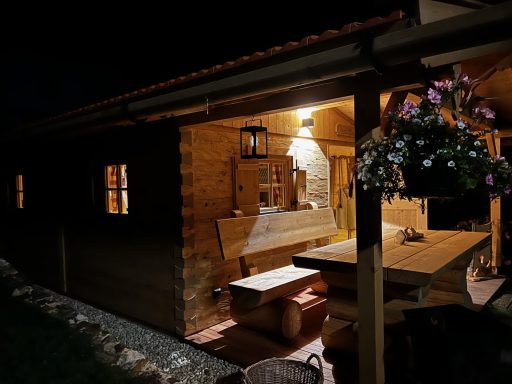 Gemütliche Holzhütte bei Nacht mit beleuchtetem Innenraum und Tisch im Freien.