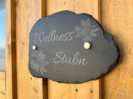 Schiefer-Schild mit der Aufschrift "Wellness Stubn" und Blumenmuster.